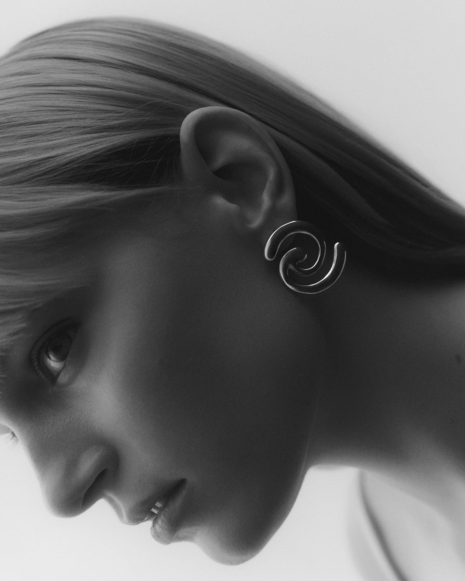 A close-up side profile of a woman wearing a silver spiral drop earring against a light gray background.