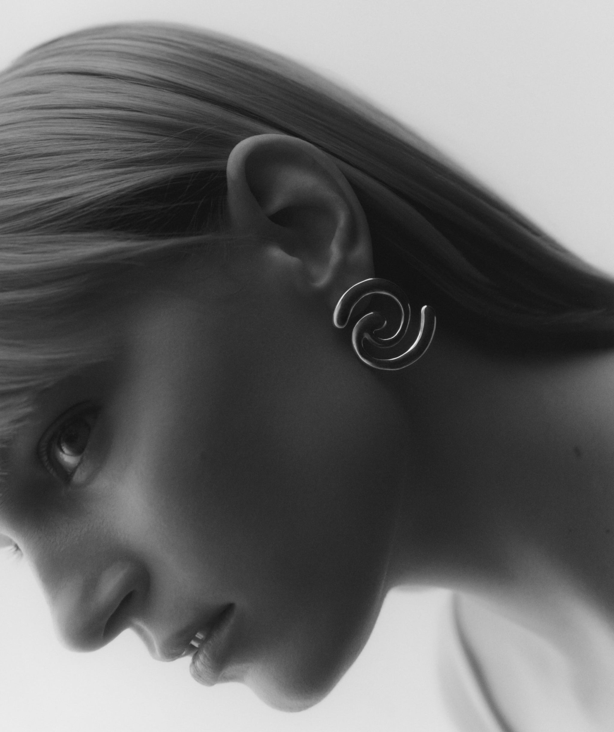 A close-up side profile of a woman wearing a silver spiral drop earring against a light gray background.