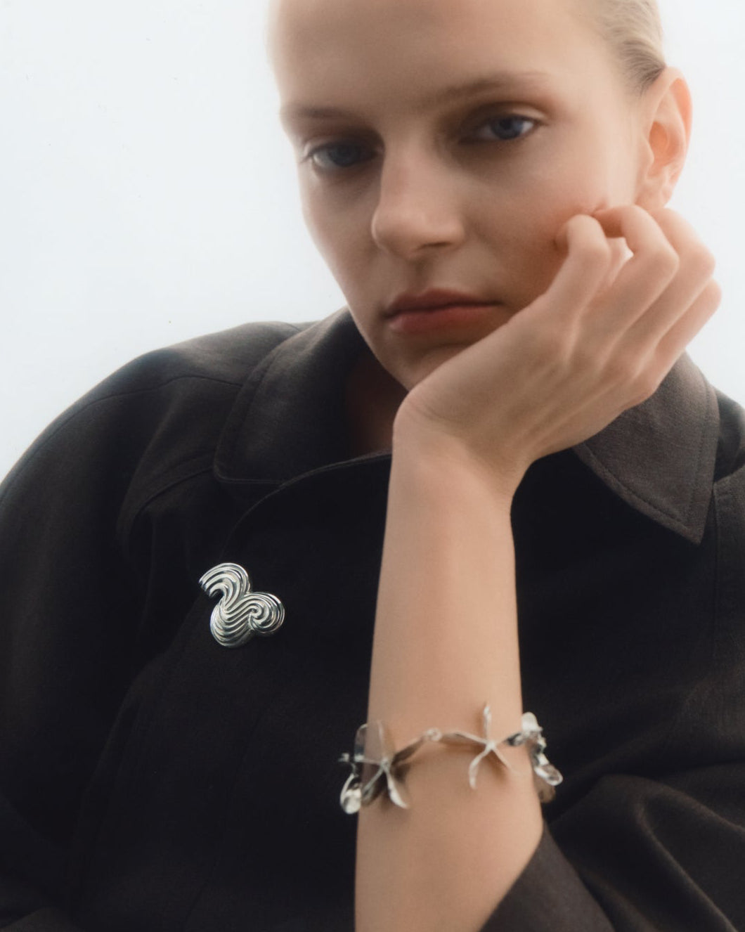 A close-up portrait of a model wearing a black jacket with a silver brooch on the chest and a silver bracelet on the wrist.