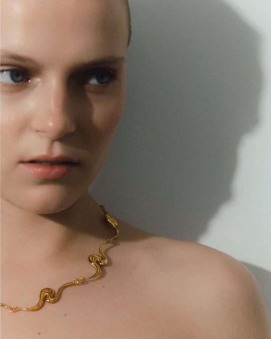 A close-up lifestyle shot of a model wearing a gold necklace against a neutral gray background with a soft shadow.
