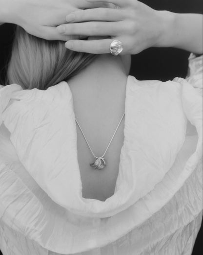 A black and white close-up of a person from behind wearing a delicate pendant necklace and a large ring on one finger, with hands covering the head area.