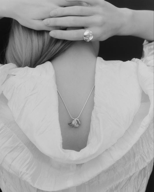 A black and white close-up of a person from behind wearing a delicate pendant necklace and a large ring on one finger, with hands covering the head area.