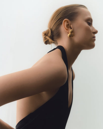 A woman in profile wearing a black sleeveless top and a textured gold earring, photographed against a clean off-white background.