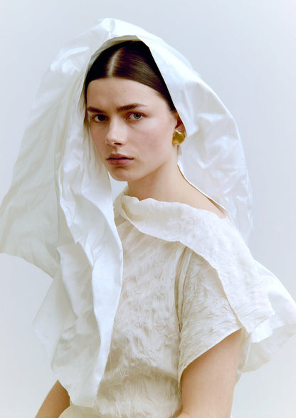 Woman wearing a white headscarf and gold earrings against a plain background