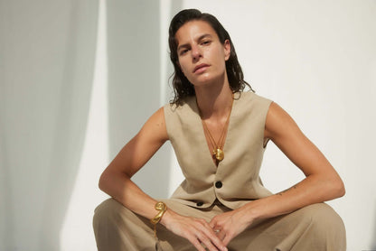 Model sitting in a beige suit, wearing a large, sculptural gold bracelet and gold necklaces