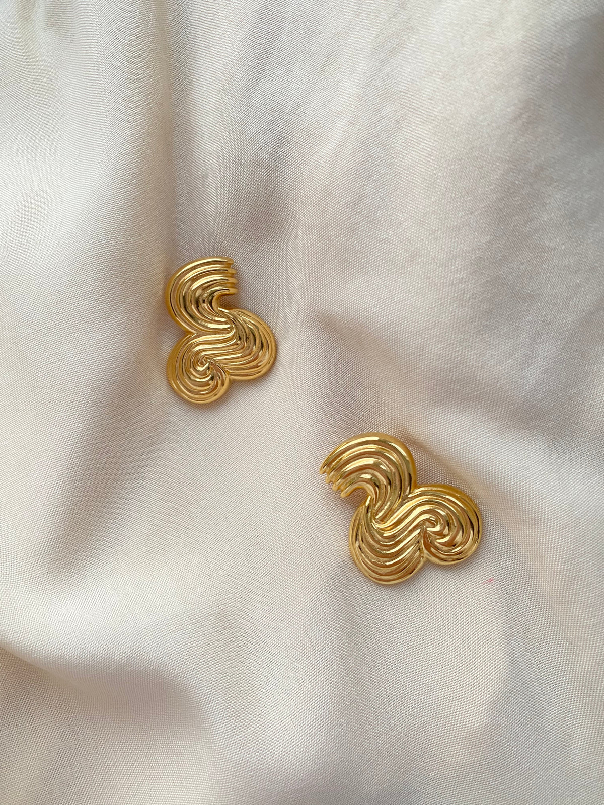 A pair of gold-toned, textured spiral stud earrings resting on a draped white fabric background.