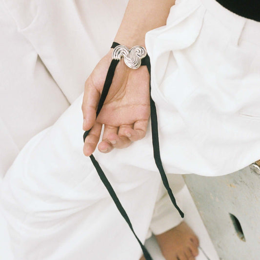 Models hand laying on his leg with white trousers, wearing silver bracelet with black silk ribbon