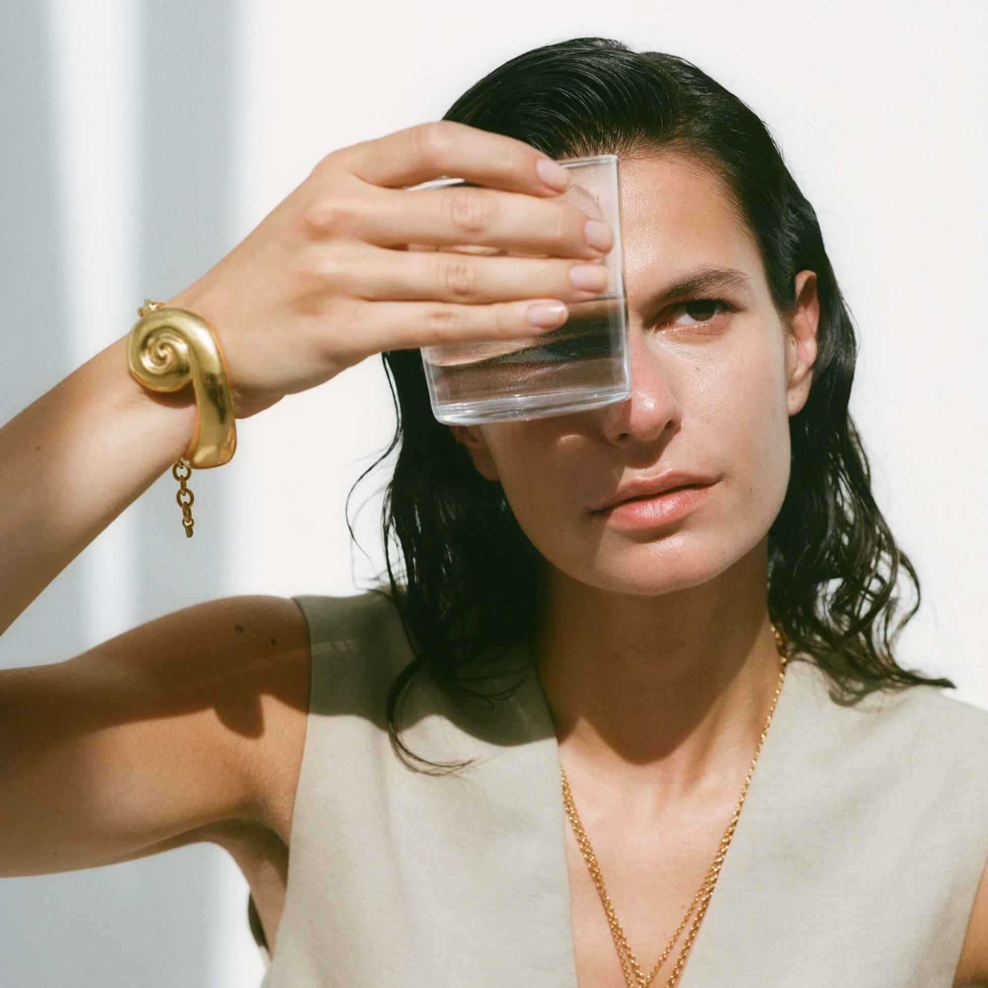 Model holding a glass of water in front of her eye, wearing a large, sculptural gold bracelet