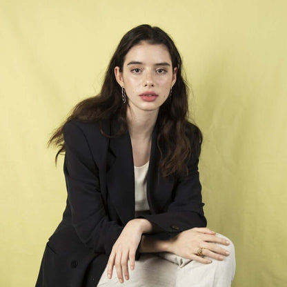 Woman sitting on a chair looking serious into the camera. Wearing a black blazer. Crossed hands and legs.